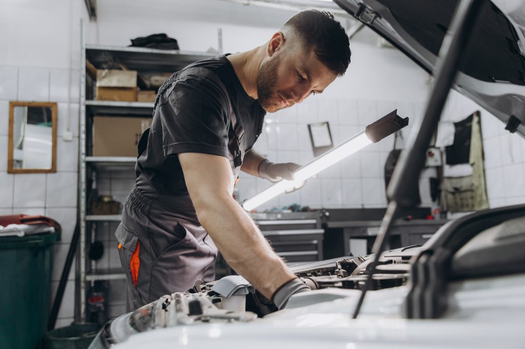 Car service, repair, maintenance and people concept - car mechanic man with lamp working at workshop